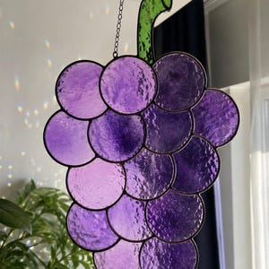 Puede incluir: Un racimo de uvas de vidriera en tonos morados, con un tallo verde. El vidrio es texturizado y las uvas están dispuestas en forma de racimo clásico. La pieza se suspende mediante una cadena, adecuada para colgar como decoración de ventana.