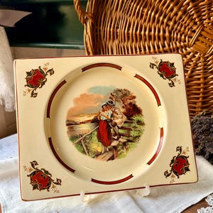 Puede incluir: Plato cuadrado de cerámica color crema con borde rojo. El plato presenta una imagen central de una pareja cerca de una masa de agua y detalles florales rojos decorativos en cada esquina.