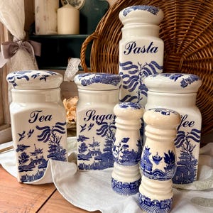May include: A set of white ceramic kitchen storage jars and salt and pepper shakers with blue willow pattern designs. The jars are labelled "Tea", "Sugar", "Pasta", and "Coffee". The set is displayed on a white cloth.