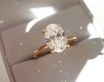 May include: A gold ring featuring a large, oval-cut, clear faceted gemstone held by four prongs. The ring is displayed inside a light-colored, open jewelry box, with sunlight creating bright reflections and sparkles on the stone and band.