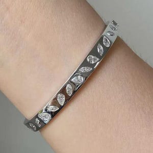 May include: A silver-colored bangle bracelet with a leaf-like pattern of clear, sparkling stones. The bracelet is worn on a wrist and reflects light, showcasing the intricate design and elegant style.