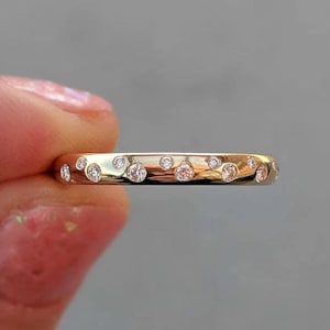 Op de afbeelding: Een gouden ring met een reeks kleine, ronde, heldere stenen die verzonken in het metalen oppervlak zijn geplaatst. De stenen zijn in een verspreid, onregelmatig patroon gerangschikt over de buitenomtrek van de gepolijste geelgouden band.