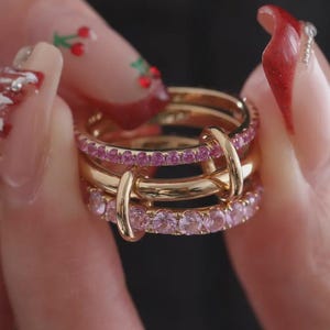 Könnte beinhalten: Ein Stapel goldener Ringe mit rosa Edelsteinen. Die Ringe werden von einer Person mit manikürten Nägeln gehalten. Die Ringe haben ein zartes Design, wobei die rosa Edelsteine in einer Reihe um das Band angeordnet sind.