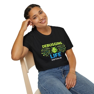 Puede incluir: Camiseta negra con el texto "DEBUGGING LIFE" en verde y azul, con un diseño de placa de circuito y un icono de error. La palabra "SKETCHLAB" está debajo. La camiseta la lleva una persona.