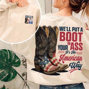 Könnte beinhalten: Cremefarbenes Sweatshirt mit einer Grafik von Cowboy-Stiefeln mit der amerikanischen Flagge und dem Text "WE'LL PUT A BOOT IN YOUR ASS It's The American Way." Eine kleinere Grafik auf der Vorderseite lautet "BROUGHT TO YOU COURTESY OF THE RED, WHITE & BLUE."