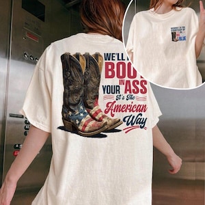 May include: Cream-colored t-shirt featuring a graphic of cowboy boots with an American flag design. The text on the shirt reads "WE'LL BOU in YOUR ASS It's The American Way." A smaller graphic on the front includes the text "BROUGHT TO YOU COURTESY OF RED, WHITE, AND BLUE."