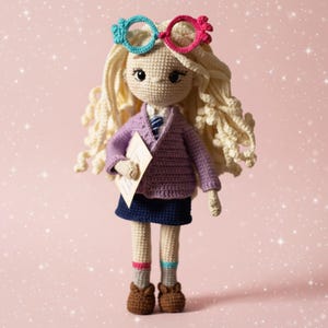 Peut inclure: Poupée au crochet faite à la main avec des cheveux blonds bouclés, portant des lunettes Spectrespecs bleues et roses, une veste violette, une jupe bleue et des chaussures marron. La poupée tient un papier et porte des chaussettes rayées.