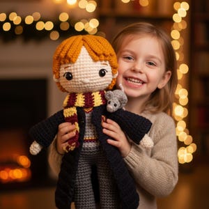May include: A crocheted doll with red hair, a scarf, and a black robe, held by a smiling child. The doll has a small gray mouse attached. The background is blurred with warm, out-of-focus lights.