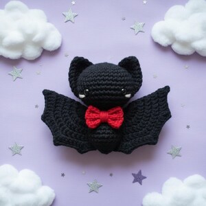 Peut inclure: Une chauve-souris noire au crochet avec un nœud papillon rouge. La chauve-souris a de grandes ailes texturées et de petits accents blancs autour des yeux. Le fond est violet clair avec des décorations de nuages et d'étoiles blanches.
