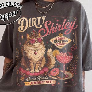 May include: A charcoal gray t-shirt featuring a graphic of a fluffy cat wearing a crown and heart-shaped glasses, sitting on a pink cushion. The text reads Dirty Shirley, A Drink Befitting Royalty, and Mama Needs A NIGHT OFF, with a cocktail.