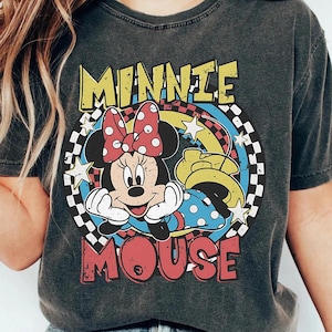 Pode incluir: T-shirt cinza escuro com um gr&aacute;fico da Minnie Mouse. O design inclui o rosto da Minnie, um la&ccedil;o vermelho com bolinhas e as palavras "MINNIE MOUSE" em amarelo e vermelho. O fundo tem um padr&atilde;o xadrez e estrelas.