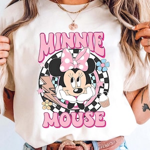 May include: A cream-coloured t-shirt with a graphic of Minnie Mouse wearing a pink bow, surrounded by flowers, stars, and the words "MINNIE MOUSE" in pink. The design is set against a black and white chequered background.