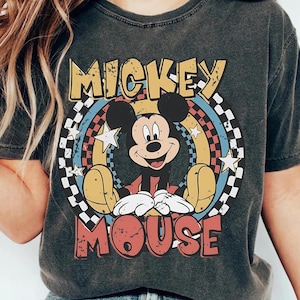 May include: Dark gray t-shirt featuring a distressed graphic of Mickey Mouse. The design includes the words "MICKEY MOUSE" in yellow and red, with a colorful rainbow and star background. The shirt has a vintage, worn look.