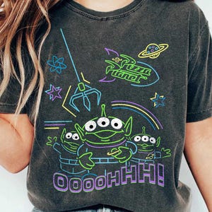 May include: Dark gray t-shirt featuring a neon graphic of the Pizza Planet aliens from Toy Story. The design includes the Pizza Planet logo, a claw, and the phrase "OOOOHHH!". The aliens are green with three eyes.