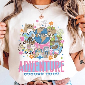 Op de afbeelding: Crèmekleurig T-shirt met een kleurrijk grafisch ontwerp. Het ontwerp bevat het woord "UNIVERSAL" op een wereldbol, omringd door illustraties van attracties en personages. Daaronder staat de tekst "ADVENTURE STARTS HERE". Het shirt wordt gedragen met een spijkerbroek en een bruine riem.