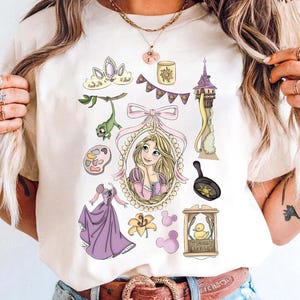 May include: Cream-colored t-shirt featuring a collection of cartoon illustrations. The design includes a princess in a pink dress, a tower, a frying pan, a crown, and other related elements. The shirt is paired with blue jeans and a brown belt.