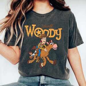 May include: Dark gray t-shirt featuring a cartoon image of Sheriff Woody riding a horse. The text "Woody" is in orange block letters with a star in the center. The shirt is a crew neck style.