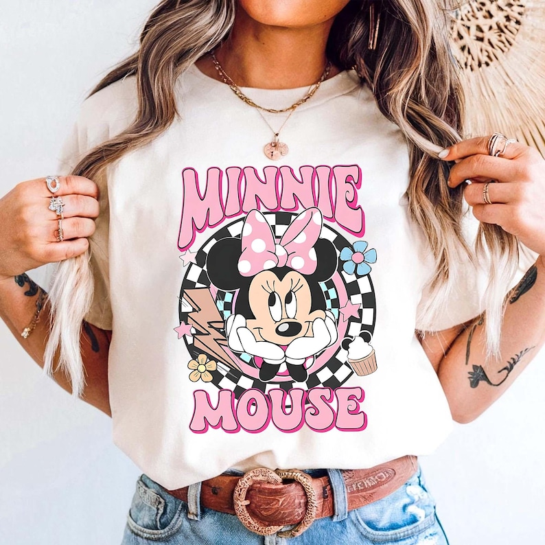 May include: A cream-coloured t-shirt with a graphic of Minnie Mouse wearing a pink bow, surrounded by flowers, stars, and the words "MINNIE MOUSE" in pink. The design is set against a black and white chequered background.