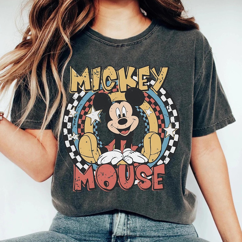 May include: Dark gray t-shirt featuring a distressed graphic of Mickey Mouse. The design includes the words "MICKEY MOUSE" in yellow and red, with a colorful rainbow and star background. The shirt has a vintage, worn look.