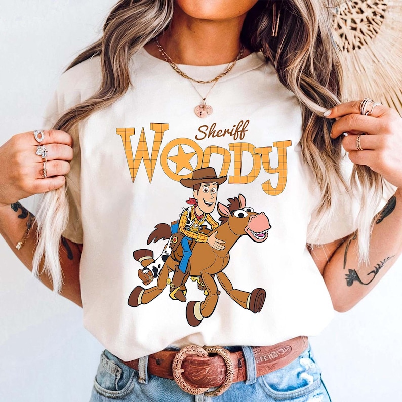May include: Cream-colored t-shirt featuring a cartoon illustration of Sheriff Woody riding a horse. The text "Sheriff Woody" is printed above the illustration in orange and yellow. The shirt has short sleeves and a crew neck.