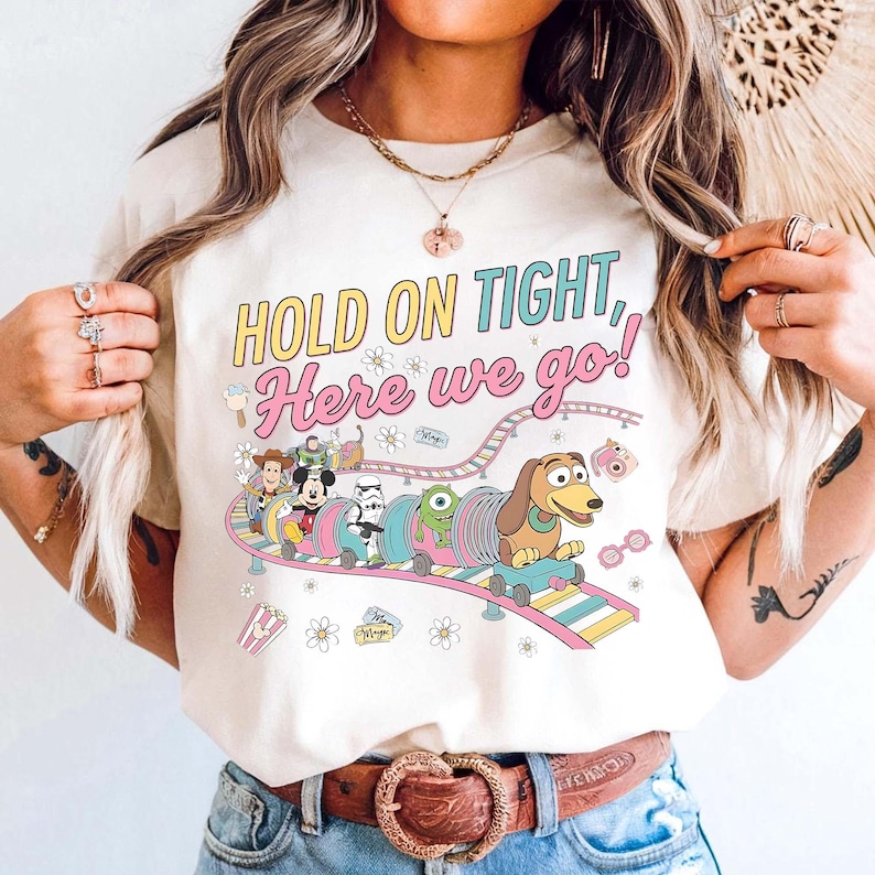Puede incluir: Camiseta de color crema con un gr&aacute;fico colorido de una monta&ntilde;a rusa de dibujos animados con personajes de Disney. El texto "HOLD ON TIGHT, Here we go!" est&aacute; impreso encima de la monta&ntilde;a rusa. La camiseta tiene cuello redondo y mangas cortas.