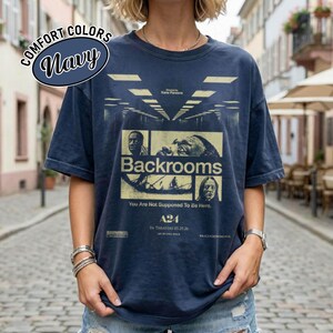 Może przedstawiać: Granatowa koszulka z kr&oacute;tkim rękawem z nadrukiem graficznym plakatu filmowego Backrooms. Projekt zawiera obrazy w odcieniach ż&oacute;łci oraz tekst Backrooms, You Are Not Supposed To Be Here, A24 i In Theaters 05.29.26 na przedniej stronie.