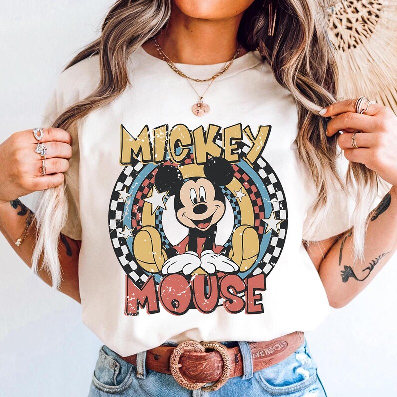 May include: Cream-colored t-shirt featuring a retro design of Mickey Mouse. The graphic includes the words "Mickey Mouse" in a distressed font, with a circular design of stars, rainbows, and a checkered pattern. The shirt has short sleeves.