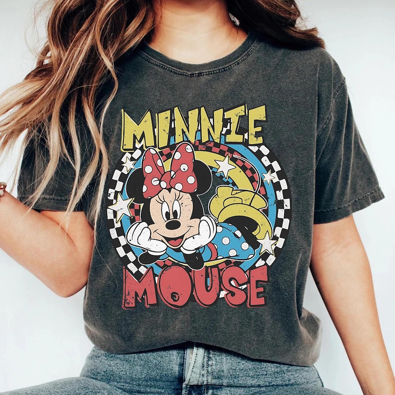 Pode incluir: T-shirt cinza escuro com um gr&aacute;fico da Minnie Mouse. O design inclui o rosto da Minnie, um la&ccedil;o vermelho com bolinhas e as palavras "MINNIE MOUSE" em amarelo e vermelho. O fundo tem um padr&atilde;o xadrez e estrelas.