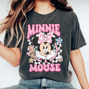 May include: Dark grey t-shirt with a graphic of Minnie Mouse wearing a pink polka dot bow, and the words "MINNIE MOUSE" in pink. The design includes flowers, a lightning bolt, and a cupcake.
