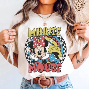 Pode incluir: Camiseta creme com um gr&aacute;fico da Minnie Mouse com um la&ccedil;o vermelho de bolinhas, rodeada pelas palavras "MINNIE MOUSE" em amarelo e vermelho. O design inclui uma borda quadriculada e detalhes de arco-&iacute;ris.