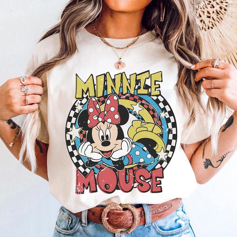 Pode incluir: Camiseta creme com um gr&aacute;fico da Minnie Mouse com um la&ccedil;o vermelho de bolinhas, rodeada pelas palavras "MINNIE MOUSE" em amarelo e vermelho. O design inclui uma borda quadriculada e detalhes de arco-&iacute;ris.