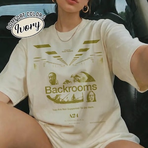 Może przedstawiać: T-shirt z kr&oacute;tkim rękawem w kolorze kości słoniowej z zielonym nadrukiem graficznym z napisem Backrooms, You Are Not Supposed To Be Here, A24 oraz In Theaters 05.29.26. Wz&oacute;r zawiera stylizowane ilustracje twarzy oraz perspektywę korytarza.