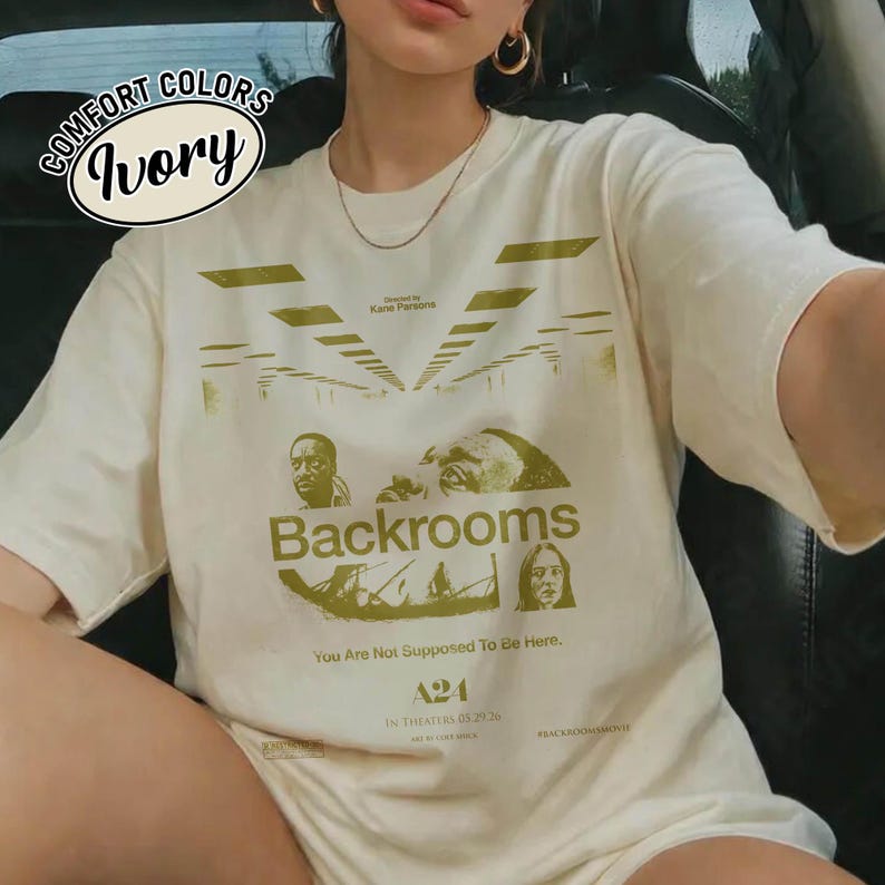 Może przedstawiać: T-shirt z kr&oacute;tkim rękawem w kolorze kości słoniowej z zielonym nadrukiem graficznym z napisem Backrooms, You Are Not Supposed To Be Here, A24 oraz In Theaters 05.29.26. Wz&oacute;r zawiera stylizowane ilustracje twarzy oraz perspektywę korytarza.