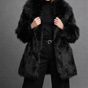 May include: A black faux fur coat with a thick, voluminous collar and long sleeves. The coat features a dense, shaggy texture throughout, extending to the mid-thigh. It is worn over a black turtleneck sweater and black trousers with a belt.