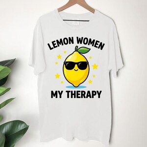 May include: White t-shirt with the text "LEMON WOMEN MY THERAPY". The shirt features a cartoon lemon wearing sunglasses, surrounded by yellow stars. The lemon is yellow with a green leaf.