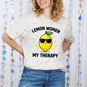 May include: A light beige t-shirt with the text "Lemon Women My Therapy" and a cartoon lemon wearing sunglasses. The shirt has a crew neck and short sleeves. The background has a star pattern.