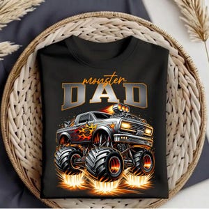 Pode incluir: Camiseta preta com um design de monster truck e o texto "monster DAD" em laranja e cinza. O design apresenta um monster truck detalhado com chamas e pneus grandes.