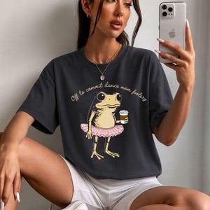 Puede incluir: Camiseta gris oscuro con una rana de dibujos animados con tutú rosa sosteniendo una taza de café. El texto "Off to commit dance mom foolery" está encima de la rana. La rana es de color marrón claro y el tutú es rosa.