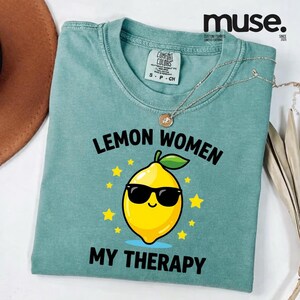 May include: A teal Comfort Colors t-shirt with the text "LEMON WOMEN MY THERAPY" and a cartoon lemon wearing sunglasses. The shirt has a crew neck and short sleeves. A silver necklace with a pendant is draped on the shirt.