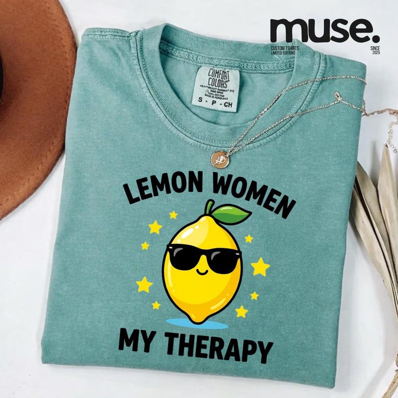 May include: A teal Comfort Colors t-shirt with the text "LEMON WOMEN MY THERAPY" and a cartoon lemon wearing sunglasses. The shirt has a crew neck and short sleeves. A silver necklace with a pendant is draped on the shirt.