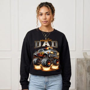 Pode incluir: Camisola preta com um gr&aacute;fico de um monster truck com chamas e o texto "monster DAD" em prata e ouro. O cami&atilde;o tem pneus grandes e emite chamas. A camisola &eacute; usada por uma pessoa.