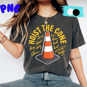 May include: Dark gray t-shirt with the text "HOIST THE CONE" in yellow, featuring an orange and white traffic cone graphic. Additional text surrounds the cone. The shirt is a casual wear item.
