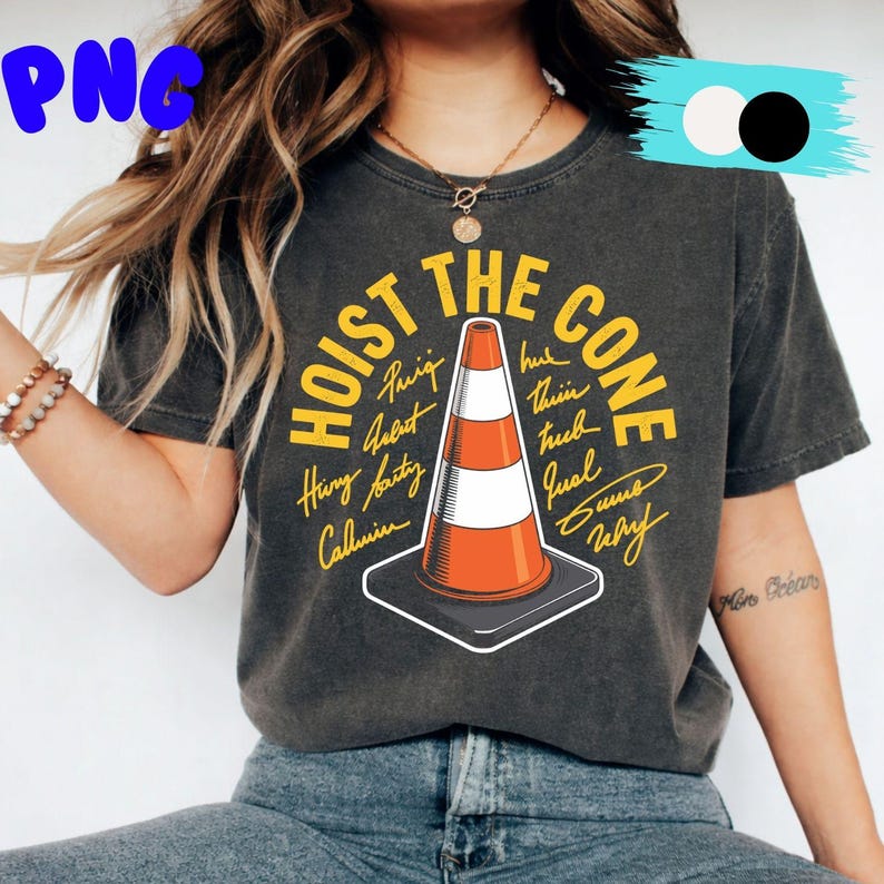 May include: Dark gray t-shirt with the text "HOIST THE CONE" in yellow, featuring an orange and white traffic cone graphic. Additional text surrounds the cone. The shirt is a casual wear item.