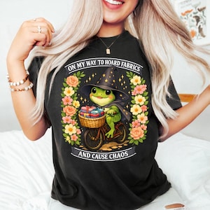 Op de afbeelding: Zwart t-shirt met een illustratie van een groene kikker met een heksenhoed en cape, fietsend met een mand vol stoffen. De tekst luidt: "ON MY WAY TO HOARD FABRICS AND CAUSE CHAOS". Omringd door bloemen.