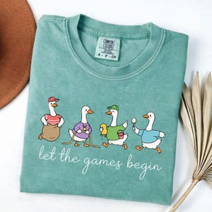 May include: A teal Comfort Colors t-shirt featuring a whimsical illustration of four geese participating in a sack race, egg and spoon race, and other games. The text "let the games begin" is written in a cursive font below the geese.