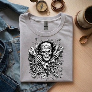 May include: A light gray t-shirt featuring a black and white graphic of a skull wearing a cap, holding wrenches, and breaking through a wall. The design includes gears, pistons, and other mechanical elements.
