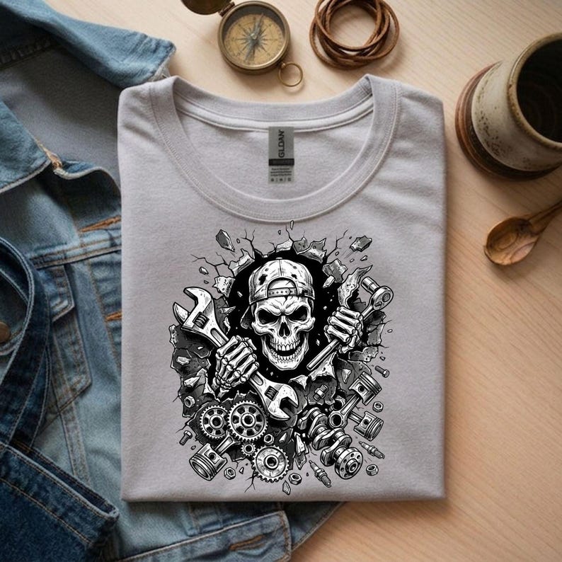 May include: A light gray t-shirt featuring a black and white graphic of a skull wearing a cap, holding wrenches, and breaking through a wall. The design includes gears, pistons, and other mechanical elements.