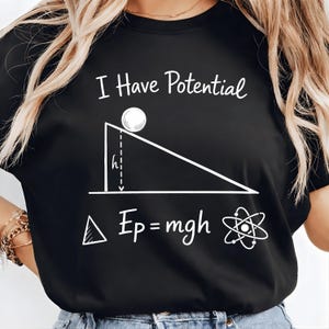 以下が含まれることがあります： 「I Have Potential」の文字と、白い物理学の図が描かれた黒いTシャツ。図には、傾斜路上のボール、数式Ep=mgh、原子の記号が含まれています。カジュアルな服装です。