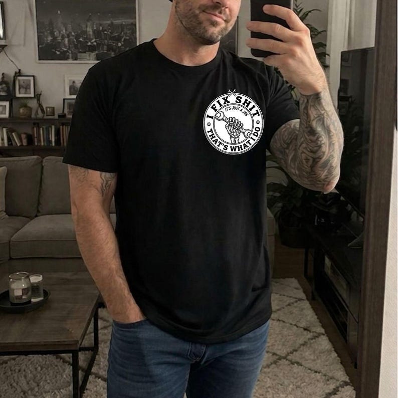 May include: Black t-shirt with a circular graphic that reads "FIX SHIT, IT'S JUST A JOB, THAT'S WHAT I DO" in white. The graphic features a wrench and a skeleton hand. The shirt is a crew neck style.