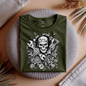 May include: An olive green t-shirt featuring a detailed graphic of a skull wearing a cap, holding wrenches, and surrounded by mechanical parts. The design is in black and white, creating a striking contrast against the shirt's color.